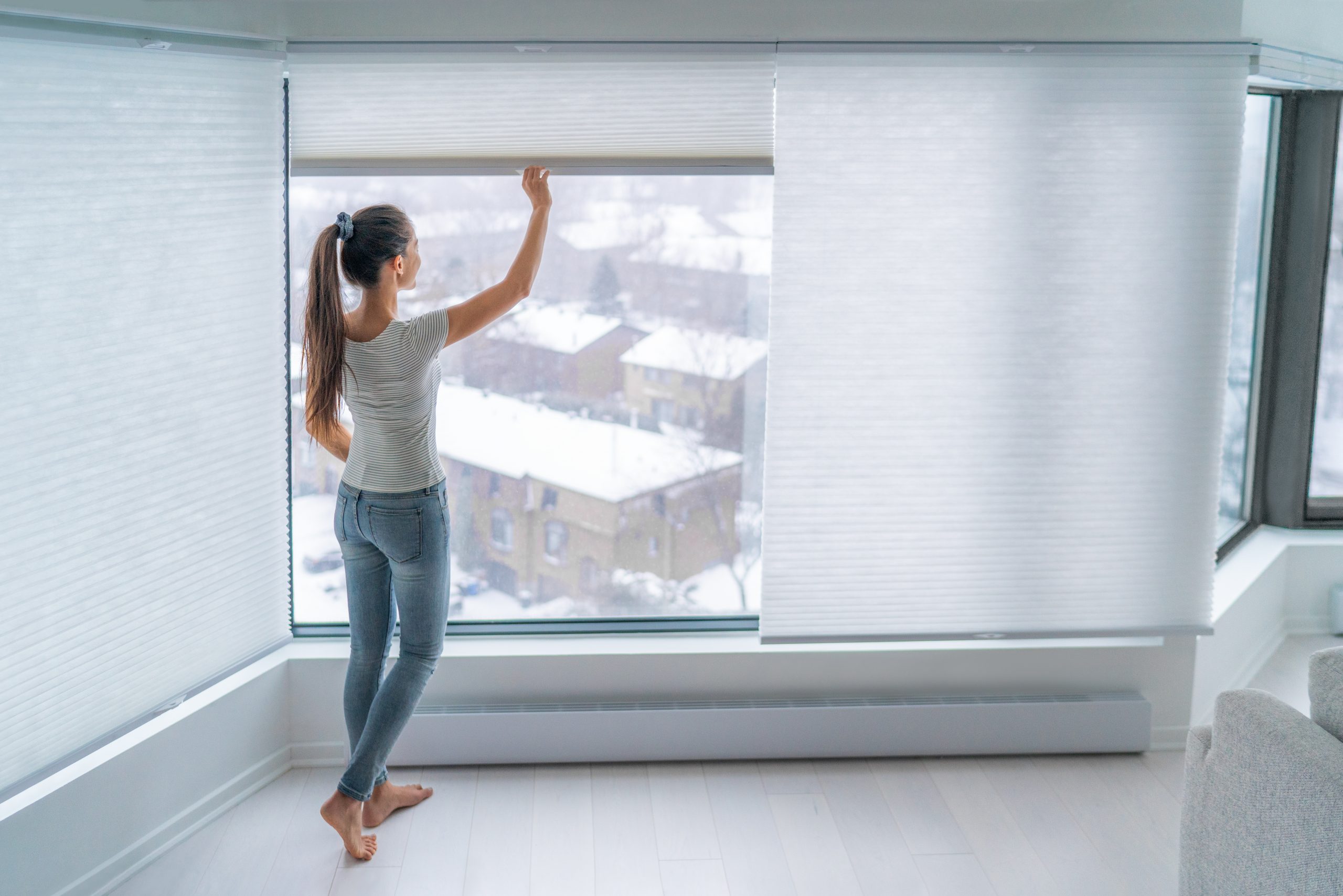 Woman closing cellular shades on apartment window keeping energy and heat indoors with honeycomb blind curtain. Cordless pleated shades in modern home living lifestyle. Interior decor design.